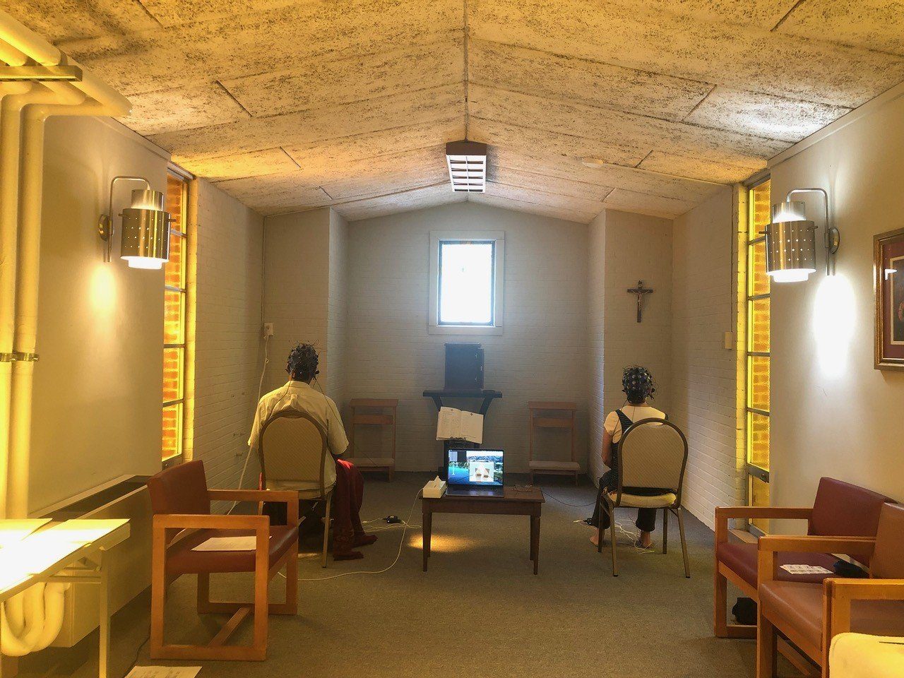 Two meditators wearing EEG caps sit in a small chapel while a laptop records their brain activity during jhāna practice.